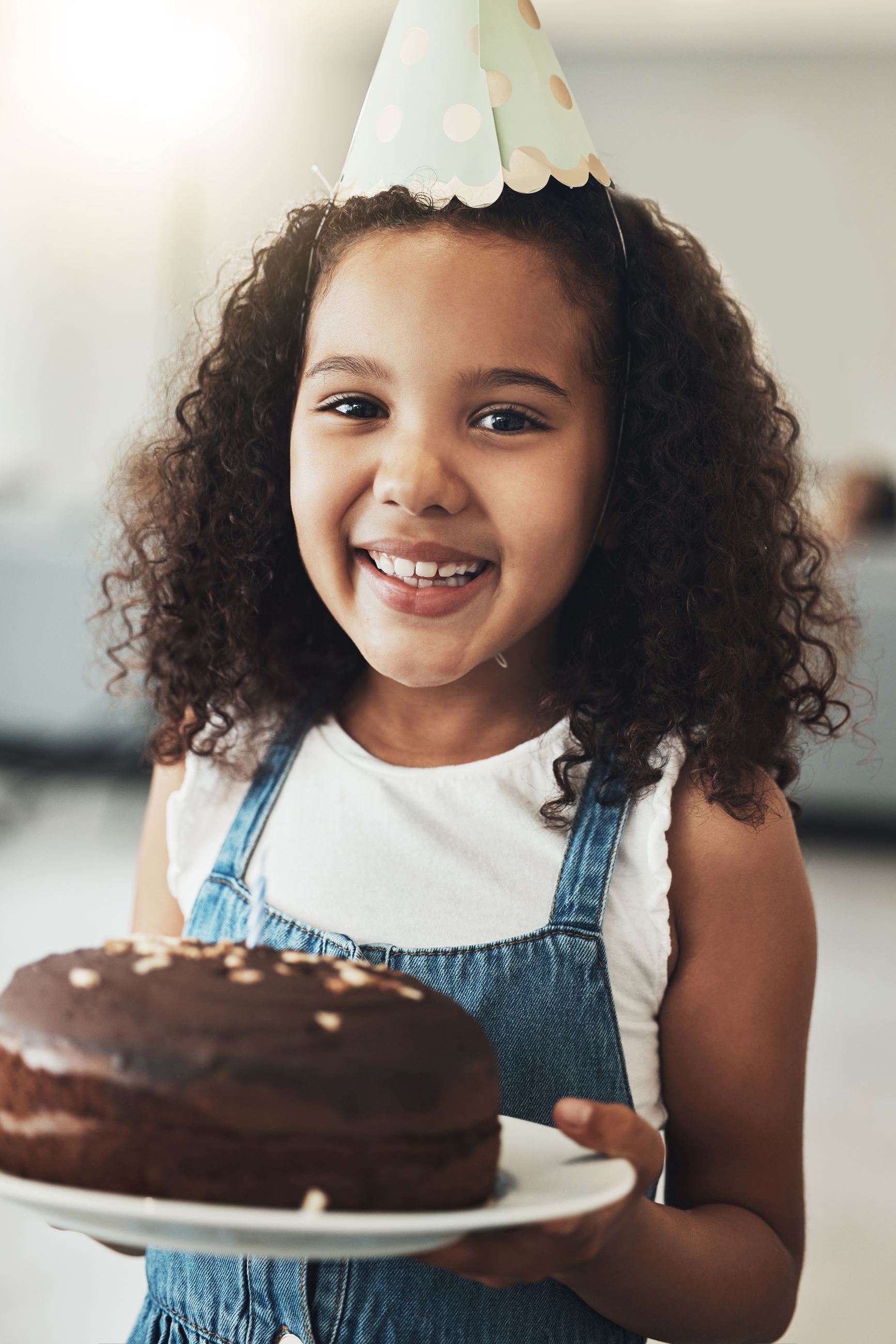 A young, happy girl in a birthday hat holds a birthday cake